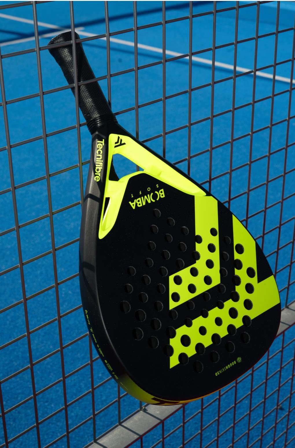 A Tecnifibre Bomba Soft Padel Racket by Tecnifibre in black and neon yellow leans against a metal fence, its fibreglass construction highlighted by the blue court backdrop.