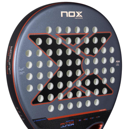 Close-up of the NOX AT10 Genius Junior By Augustin Tapia 2025 padel racket, featuring a bold black "X," circular perforations, and red details on a matte gray surface. "Junior" and "FG8" are shown on the side—ideal for young players.