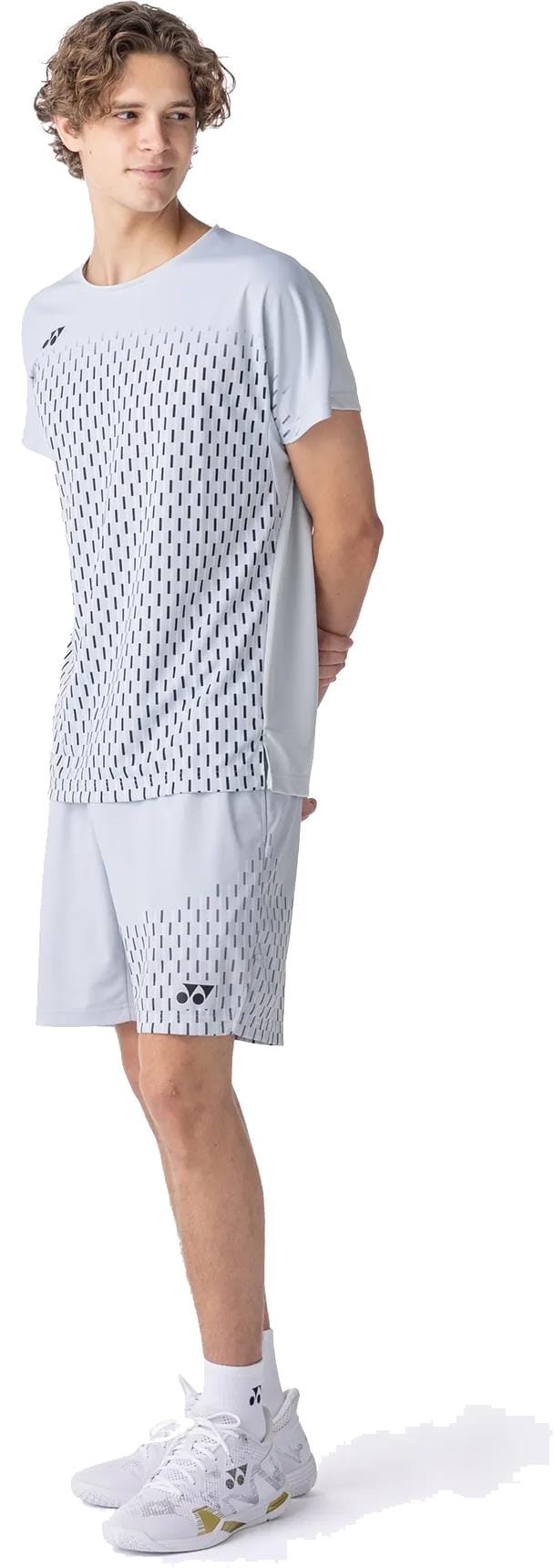 A young person with curly hair stands sideways, wearing a light gray sports t-shirt and Yonex 15241EX Knit Mens Padel Shorts in Silver Grey, paired with white athletic shoes.