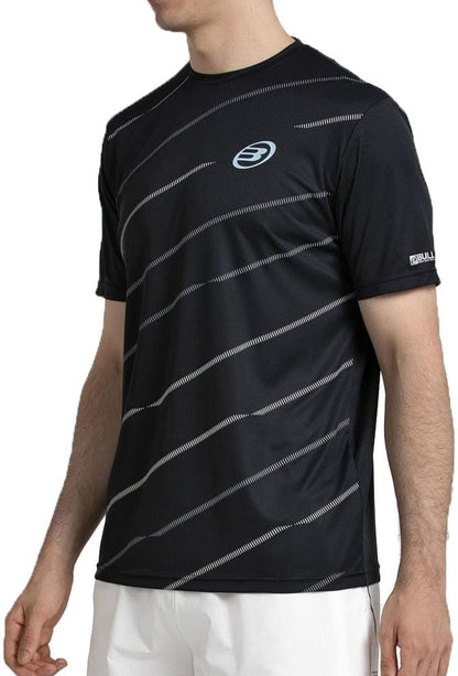 A man wears a black Bullpadel Casinas Mens Padel T-Shirt by Bullpadel, featuring diagonal white stripes and a chest logo, made from lightweight polyester with Quickerdry tech, paired with white shorts as he stands with one arm slightly bent.