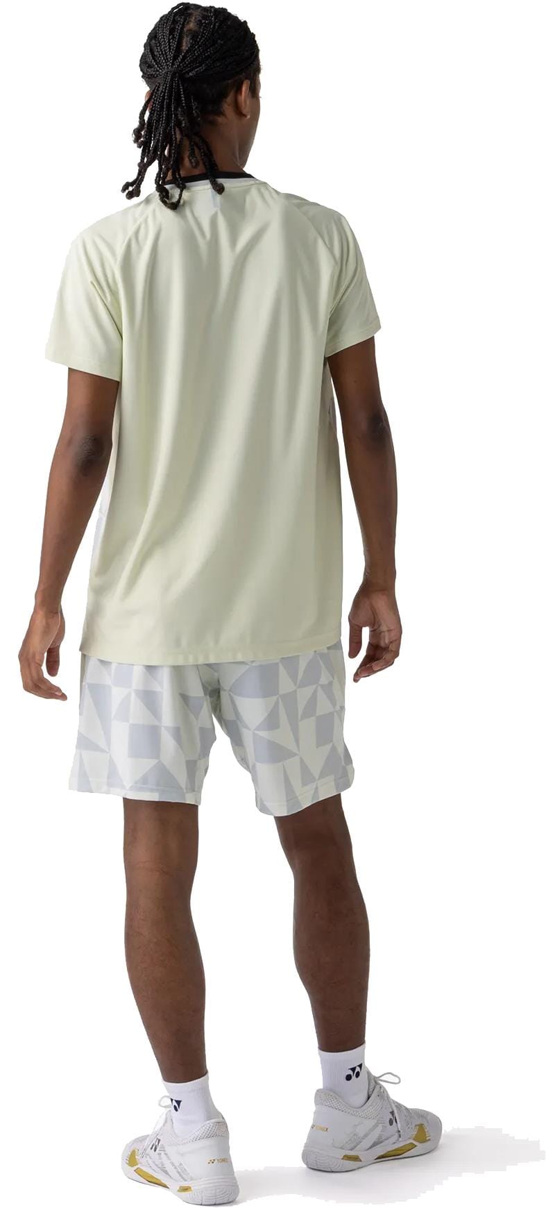 A person with braided hair wears the Yonex 10712YX Crew Neck Men's Padel T-Shirt in Champagne, along with patterned white shorts, white socks, and white sneakers, standing facing away from the camera against a white background.