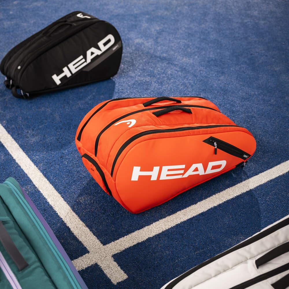 A luminous fluo orange HEAD Tour Padel Bag L is positioned on a blue tennis court amidst other sports bags. It boasts a backpack carry system and CCT+ climate control to ensure your gear remains in excellent condition.