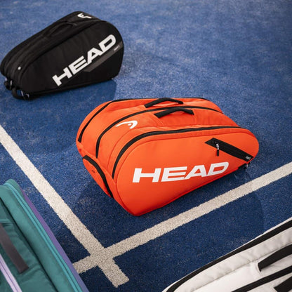 A luminous fluo orange HEAD Tour Padel Bag L is positioned on a blue tennis court amidst other sports bags. It boasts a backpack carry system and CCT+ climate control to ensure your gear remains in excellent condition.