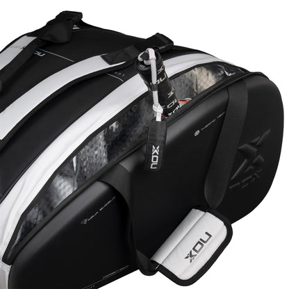 The black and white NOX Luxury Master Series Padel Racket Bag, featuring NOX branding and a textured semi-transparent side panel, is partially open to show a paddle racquet inside. It has a padded shoulder strap for comfort.