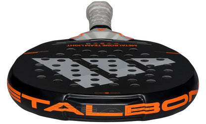 A close-up of the adidas Metalbone Team Light 2026 Padel Racket, made for advanced padel players, shows its black and orange face with perforations, "METALBONE" branding on the side, and a handle with a light-colored grip.