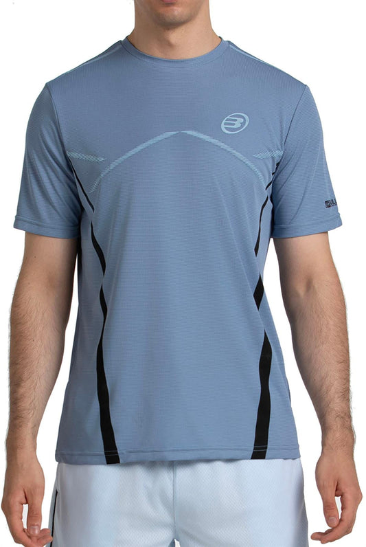 A man wears a Bullpadel Cabore Men's Padel T-Shirt in shadow blue by Bullpadel with white athletic shorts, standing against a plain white background. His face is not visible.