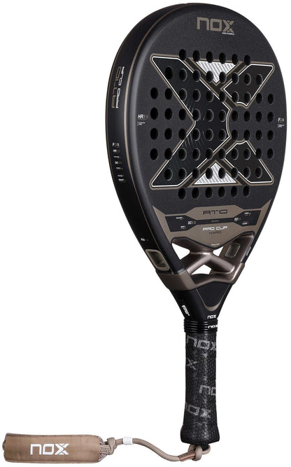 The NOX AT10 Pro Cup Hard By Agustín Tapia 2026 padel racket features a black design with a large "X", multiple face holes, textured grip, and beige wrist strap.