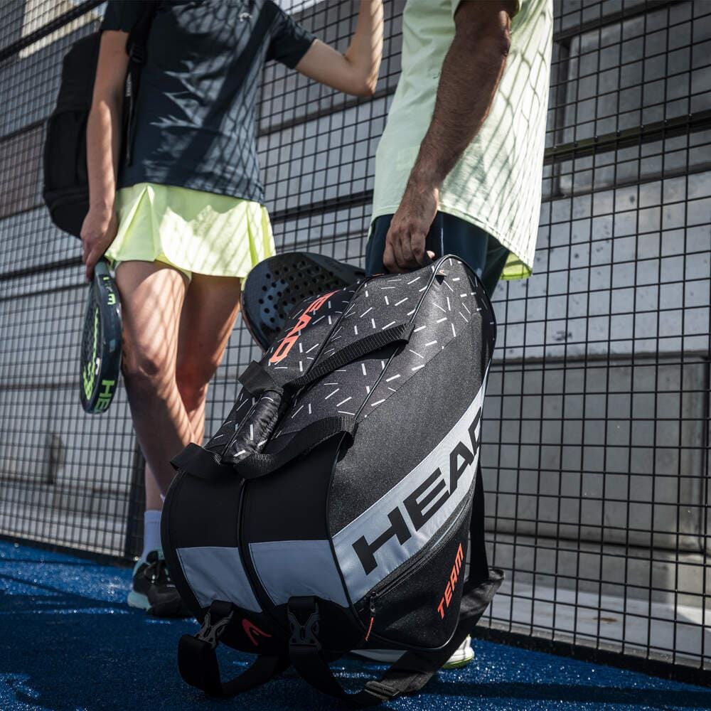 Two people stand next to each other on a sports court with a wire fence. One holds the lightweight HEAD Team Padel Bag L - BKCC, featuring multiple racket compartments. Each person holds a paddle, indicating they are about to play a racket sport.