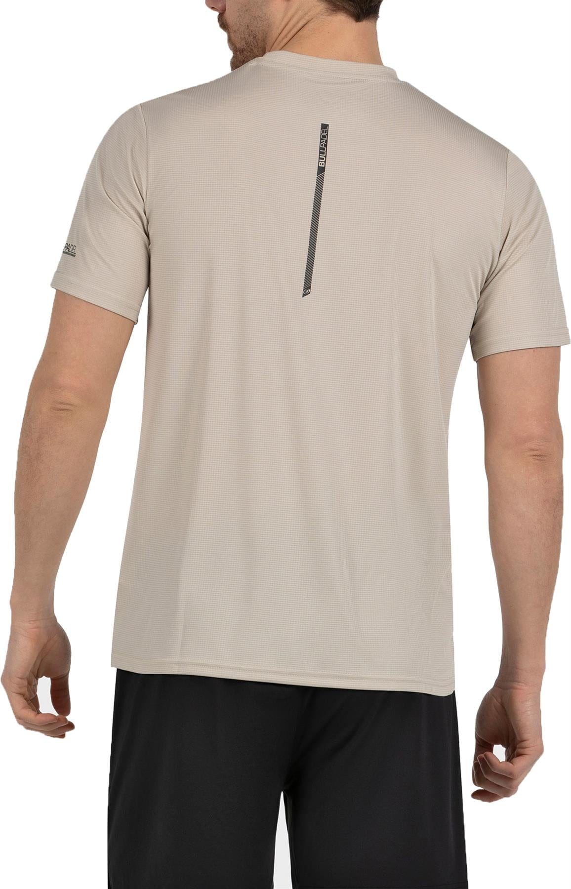 A man wearing a Bullpadel Cabore Men's Padel T-Shirt in Peanut with Quickerdry technology and black shorts, shown from the back. The shirt has a small vertical stripe with text near the upper back.