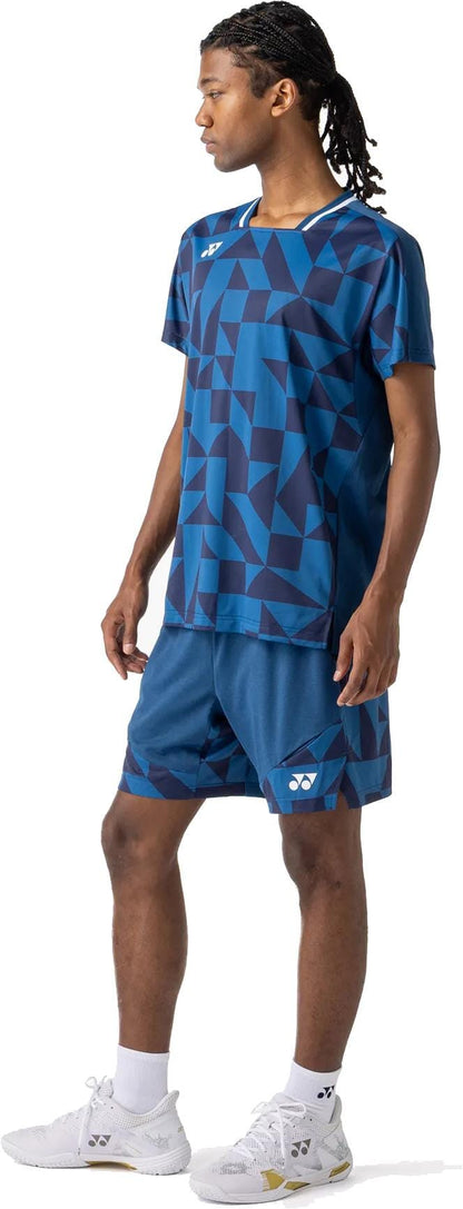 A person stands sideways in a blue geometric-patterned athletic shirt and Yonex 15240YX Knit Men's Padel Shorts – Indigo Blue by Yonex, along with white socks and sports shoes, looking left against a plain white background.