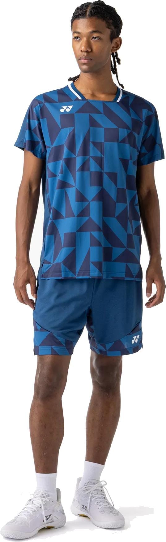 A man stands wearing the Yonex 10712YX Crew Neck Mens Padel T-Shirt in Indigo Blue, paired with matching shorts, white socks, and white athletic shoes. He looks to the side.