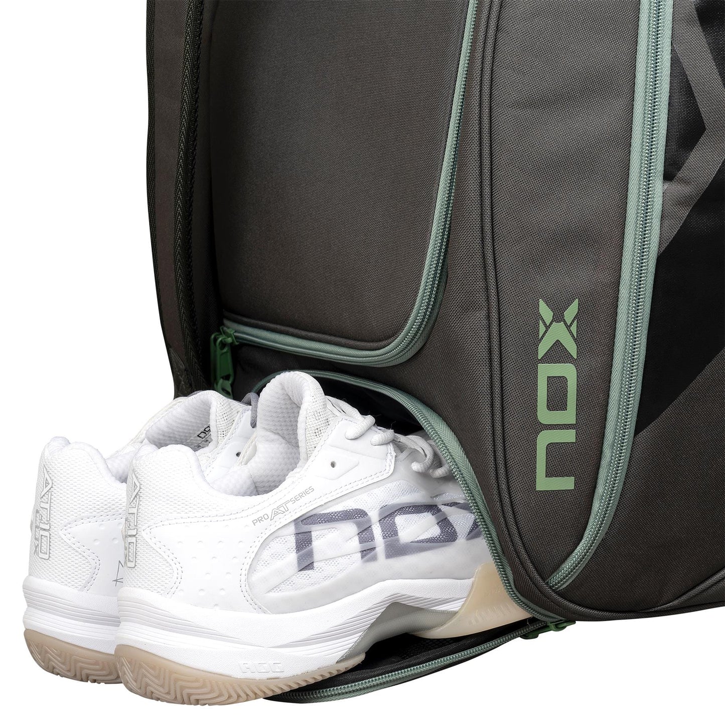 A pair of white athletic shoes is partially tucked into a zippered compartment of the NOX AT10 Competition Trolley Padel Racket Bag - Black / Green, a large wheeled padel bag with green accents and the vertical “NOX” logo on the side.