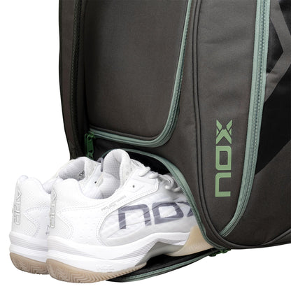 A pair of white athletic shoes is partially tucked into a zippered compartment of the NOX AT10 Competition Trolley Padel Racket Bag - Black / Green, a large wheeled padel bag with green accents and the vertical “NOX” logo on the side.