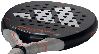 Close-up of the adidas Metalbone Team Light 2026 Padel Racket for advanced players, featuring a black surface with white and orange accents, geometric logo, “METALBONE TEAM LIGHT” text, and multiple round holes.