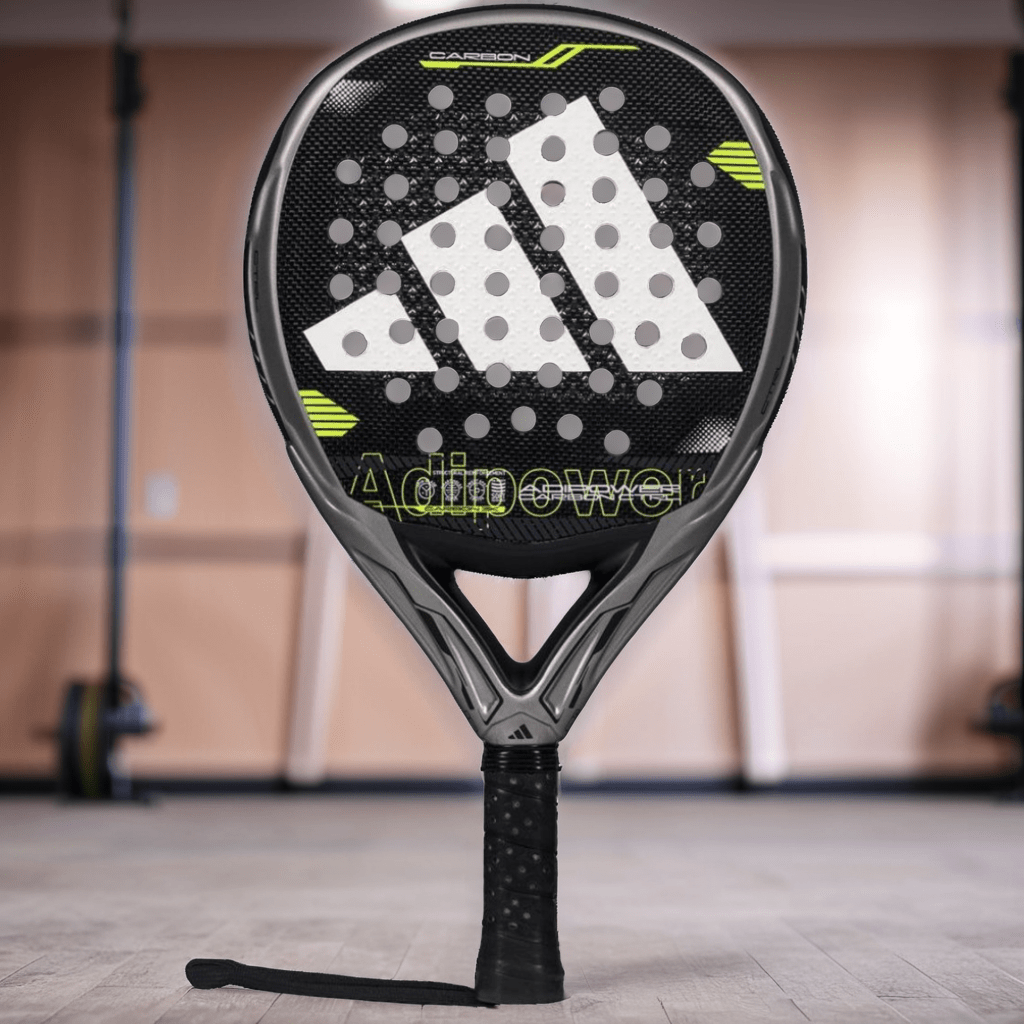 A close-up of the adidas ADIDAS Adipower Carbon CTRL 3.4 padel racket, in black and gray with yellow accents and carbon 18K construction, stands upright on a gym floor with blurred equipment in the background. "Adipower" appears on the face.