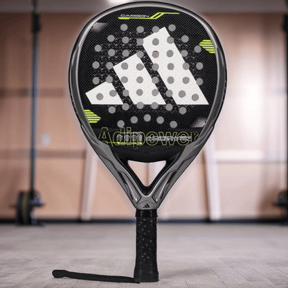 A close-up of the adidas ADIDAS Adipower Carbon CTRL 3.4 padel racket, in black and gray with yellow accents and carbon 18K construction, stands upright on a gym floor with blurred equipment in the background. "Adipower" appears on the face.