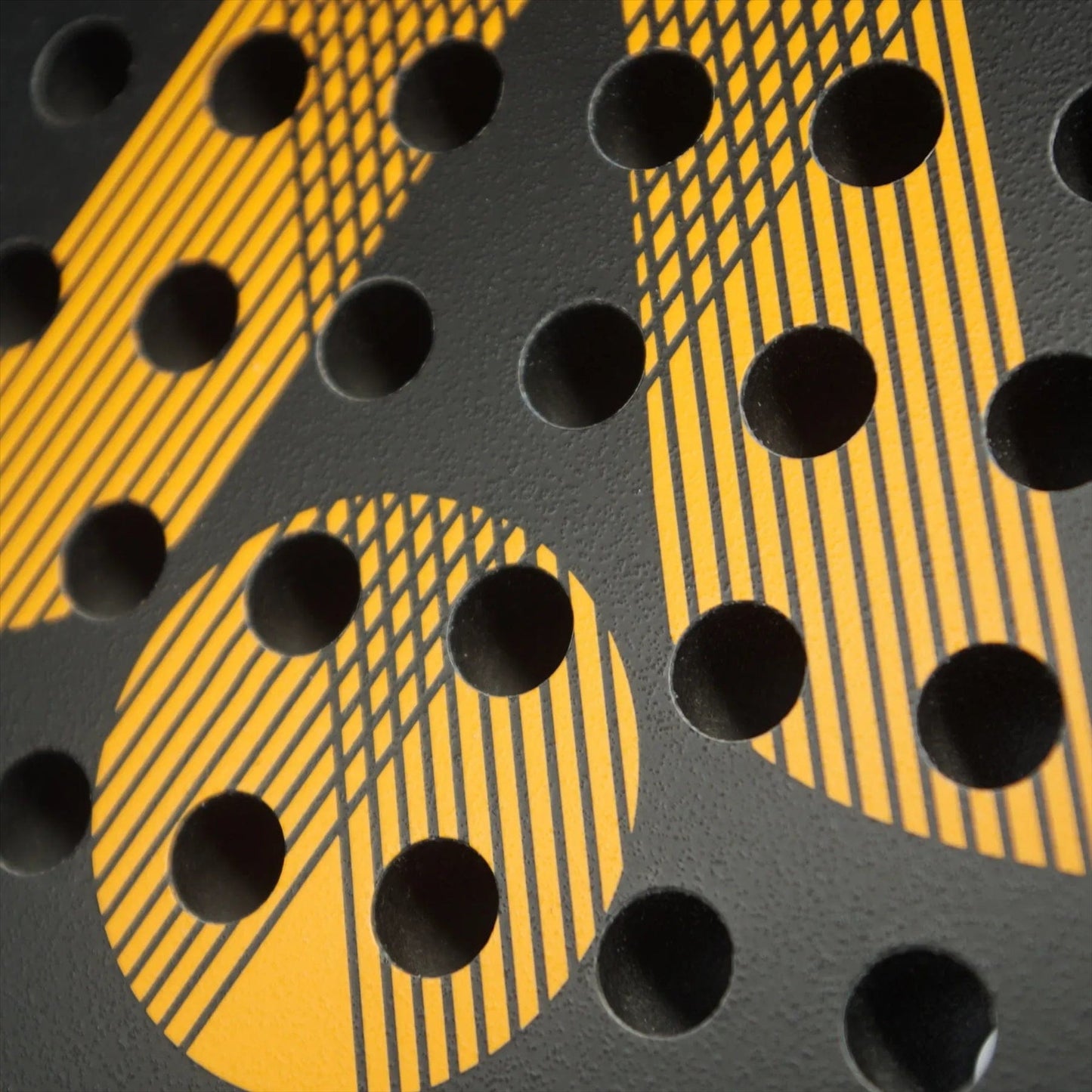Close-up of a black surface with evenly spaced circular holes and yellow diagonal stripes, forming a geometric pattern reminiscent of the Karakal Air Lite 345 Orange Padel Racket by Karakal.