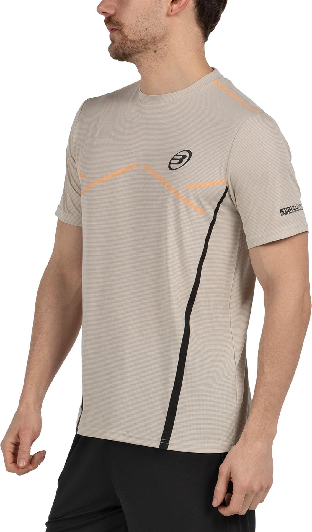 A man in a light peanut Bullpadel Cabore Men's Padel T-Shirt with Quickerdry technology and orange-black accents, paired with black shorts, stands slightly turned left with his left arm relaxed at his side.