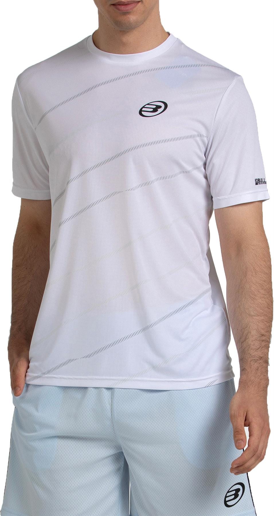 A man wears the Bullpadel Casinas Men's Padel T-Shirt in white, featuring subtle diagonal stripes, quickerdry tech, and a small black chest logo. He pairs it with light blue athletic shorts; his face is partially out of frame.