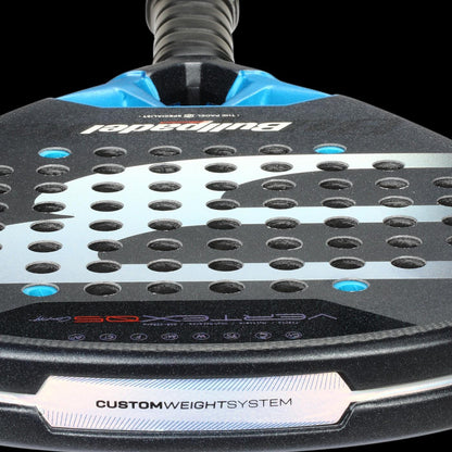 Close-up of the Bullpadel Vertex 05 Comfort 2026 Padel Racket featuring a black, blue, and silver design. Visible text includes "Bullpadel," "Vertex 05," and "CUSTOM WEIGHT SYSTEM." It has round perforations on the surface.