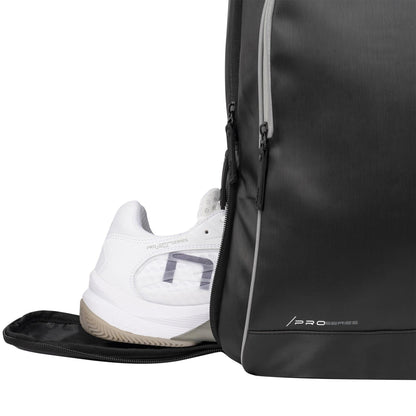 A white athletic shoe is partially tucked into a side compartment of the sleek black NOX Pro Series Padel Backpack, featuring gray zippers, "PRO SERIES" branding on the bottom corner, and ample storage for all your essentials.