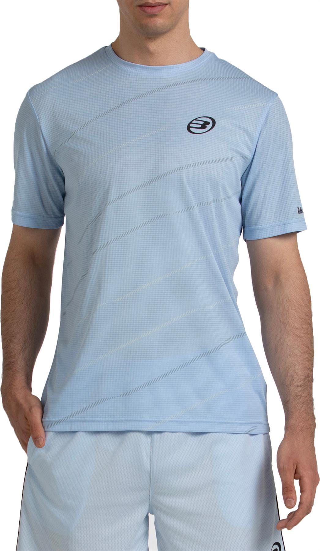 A man wears the Bullpadel Casinas Men's Padel T-Shirt in Glacier Blue, made from lightweight polyester with subtle diagonal lines, Quickerdry technology, and a small chest logo, paired with athletic shorts.