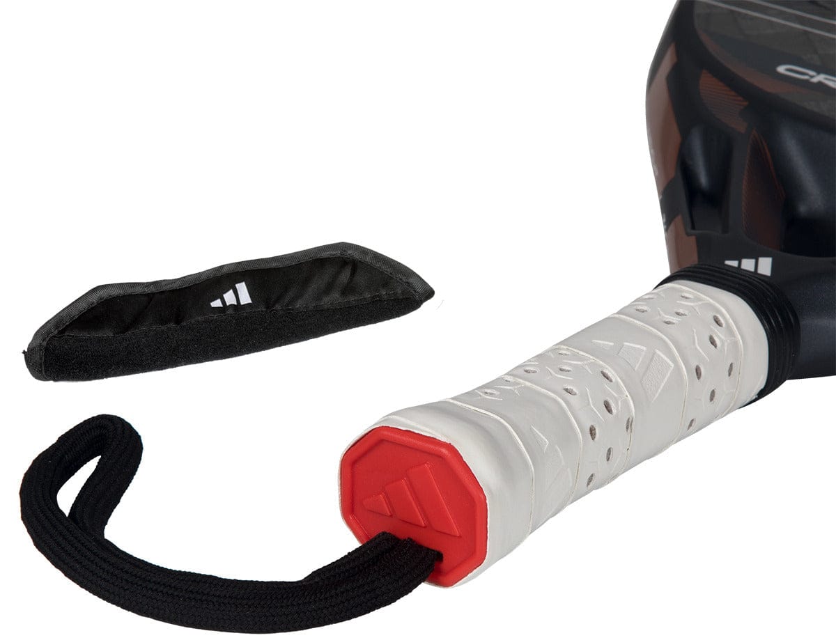 Close-up of the adidas Cross It Ctrl 2026 Padel Racket handle, featuring a white grip, red end cap, and black wrist strap. A black protective cover with a white logo lies nearby, showcasing the carbon fibre racket’s premium quality.