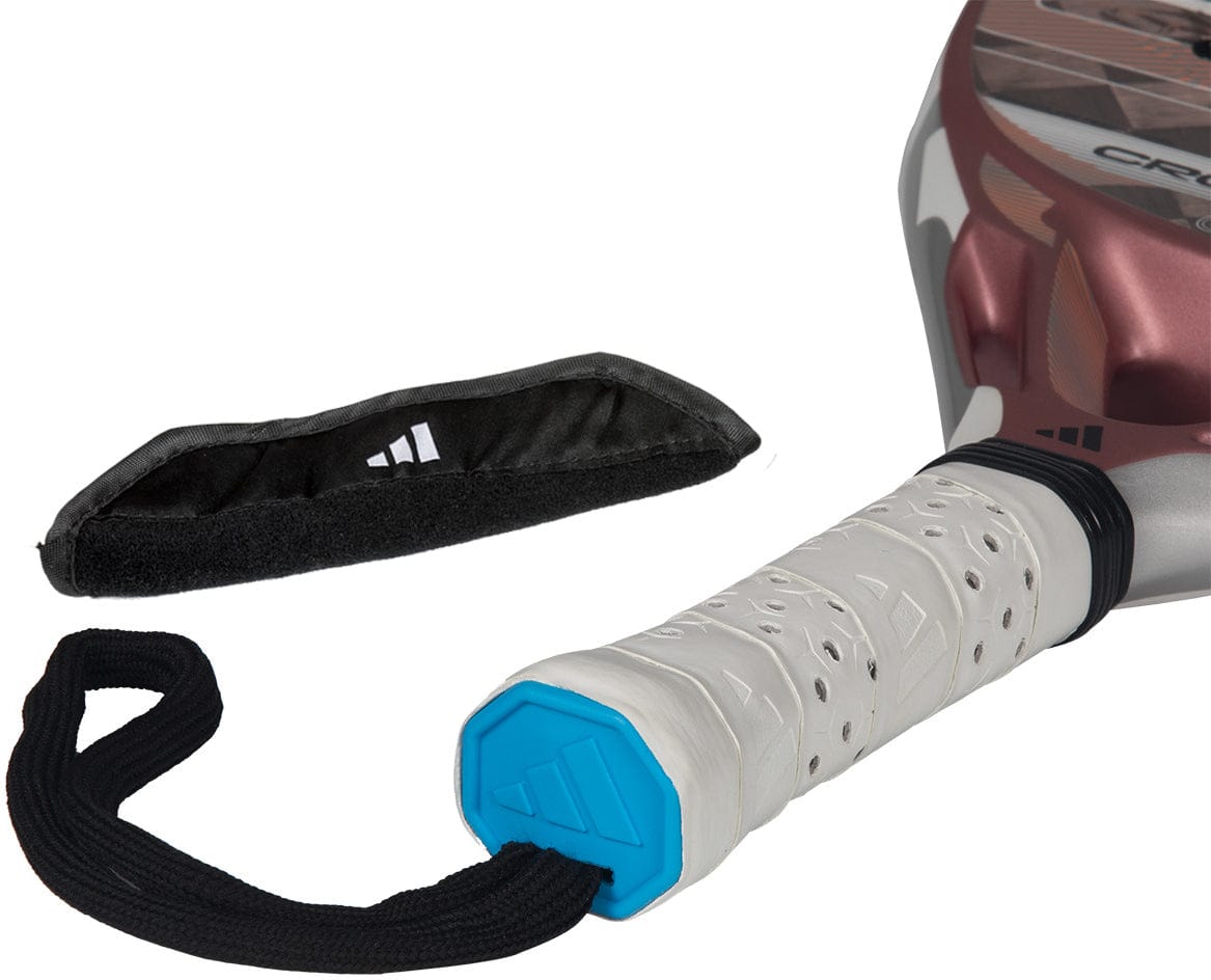 A close-up of the adidas Cross It Light 2026 Padel Racket handle features a white grip, black wrist strap, and blue end cap. Beside it lies a carbon fibre-look hook-and-loop strap with an adidas logo.