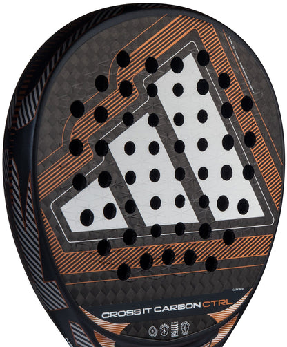 Close-up of the adidas Cross It Carbon Ctrl 2026 padel racket, featuring carbon fibre construction, orange accents, geometric design, circular holes, "CROSS IT CARBON CTRL" text, and Extra Power Grip near the bottom.