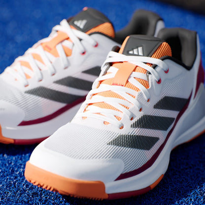 A close-up of adidas Crazyquick LS Mens Padel Shoes - White / Orange showcases their white uppers with orange and black details, black side stripes, and orange soles for grip, all placed on a textured blue surface.