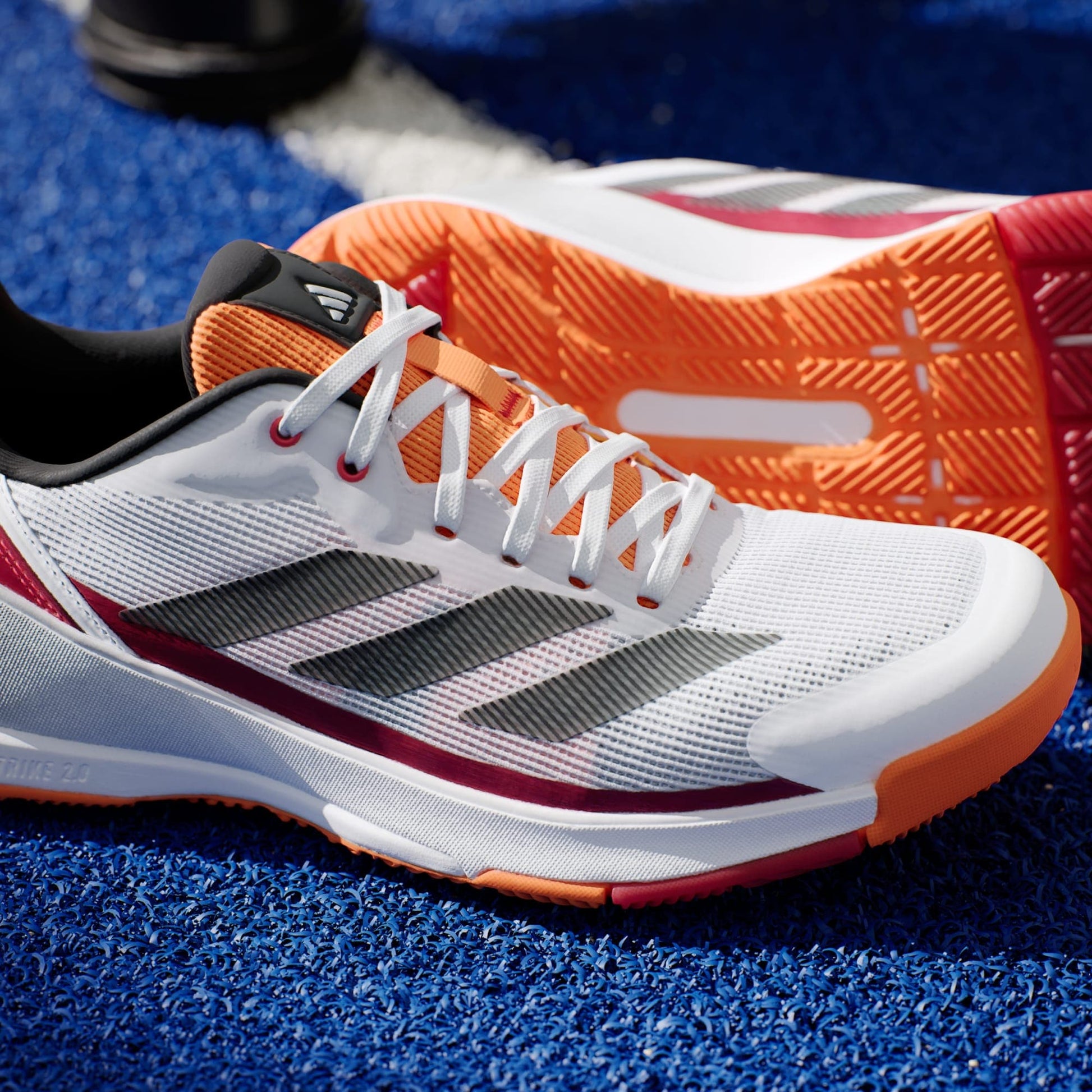 A pair of adidas Crazyquick LS Mens Padel Shoes in white and orange, with black stripes and textured orange soles for grip, rests on a blue sports surface.