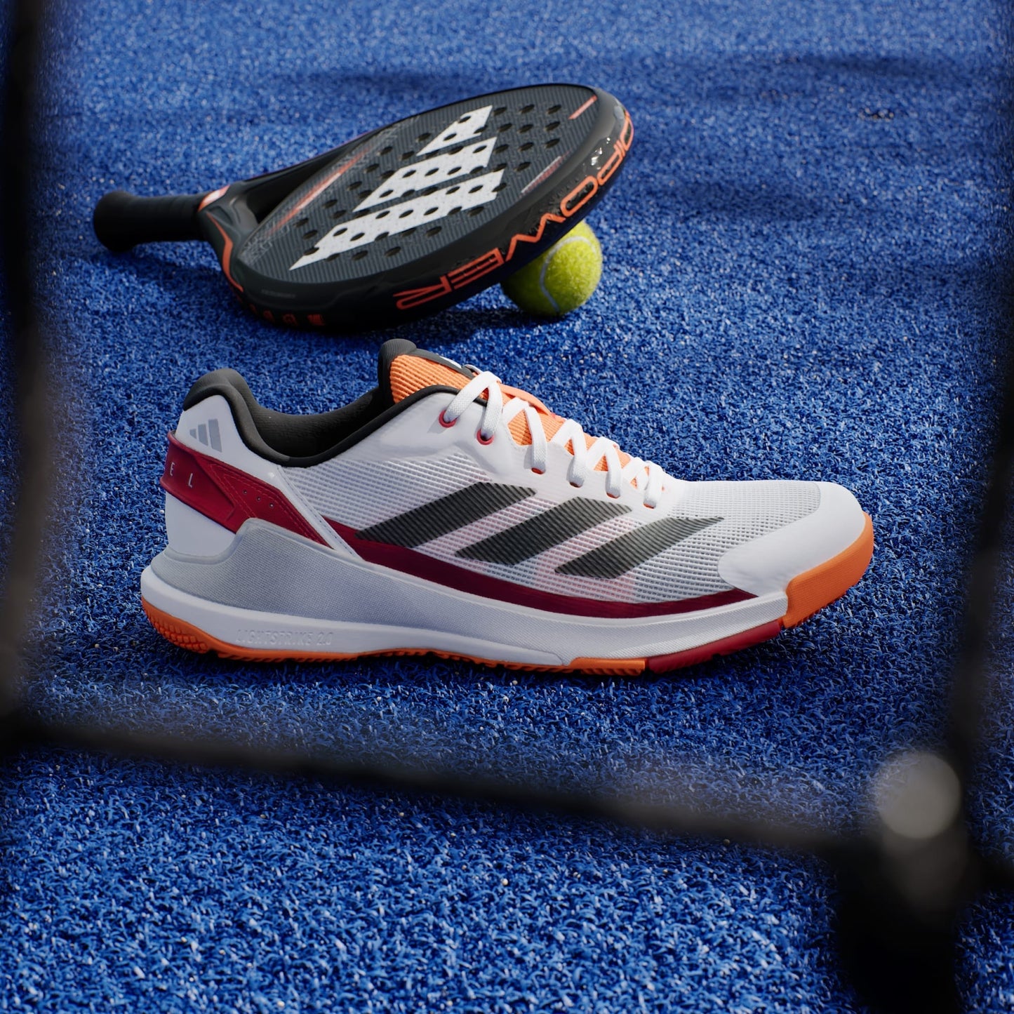 An adidas Crazyquick LS Men's Padel Shoe in white and orange rests on a blue court, beside a black paddle racket and a yellow tennis ball.