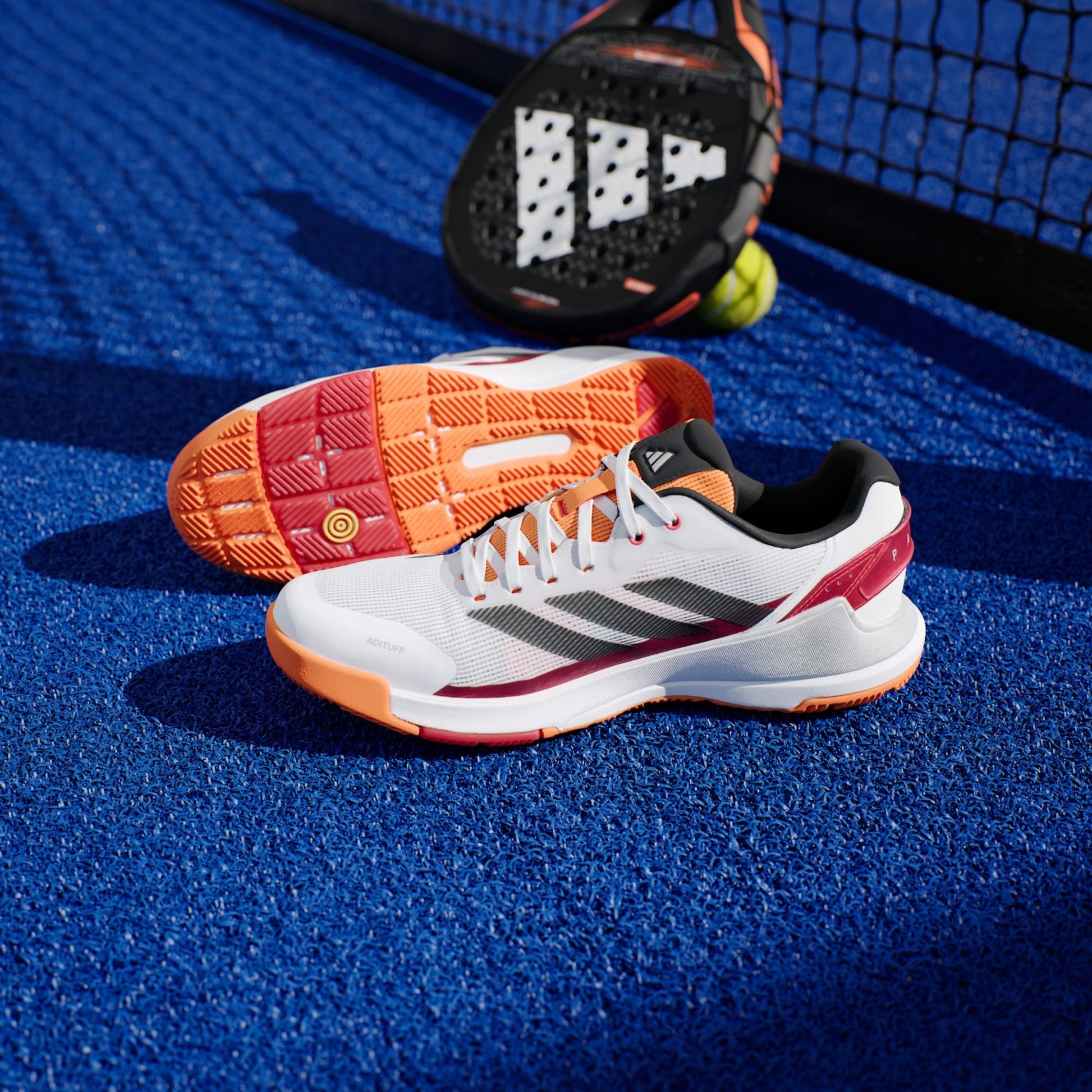 A pair of adidas Crazyquick LS Mens Padel Shoes in White/Orange rests on a bright blue tennis court, showcasing multi-directional grip, with a black padel racket and yellow balls near the net.