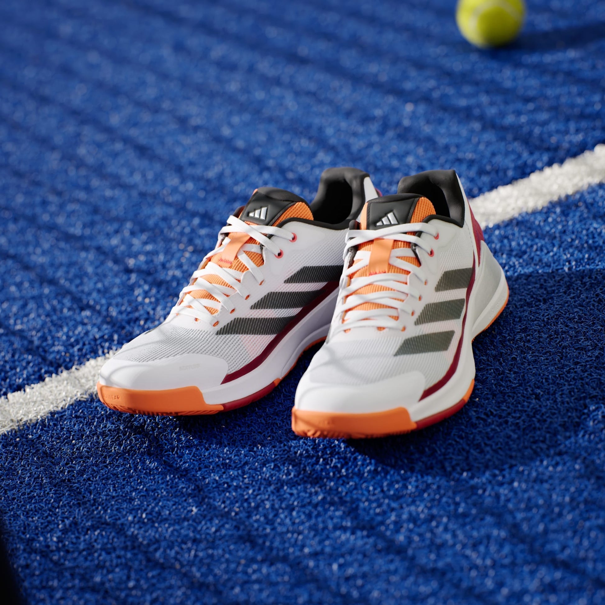 A pair of adidas Crazyquick LS Mens Padel Shoes in white and orange sit on a blue textured sports court by a white line, showcasing multi-directional grip, with a yellow tennis ball visible in the background.