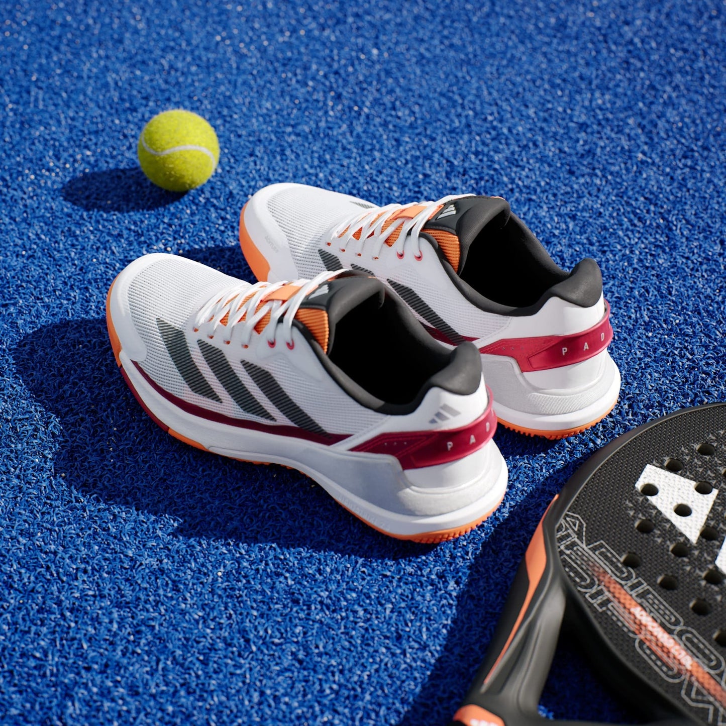 A pair of adidas Crazyquick LS Men's Padel Shoes in White/Orange are on blue turf beside a yellow tennis ball and a black paddle racket.