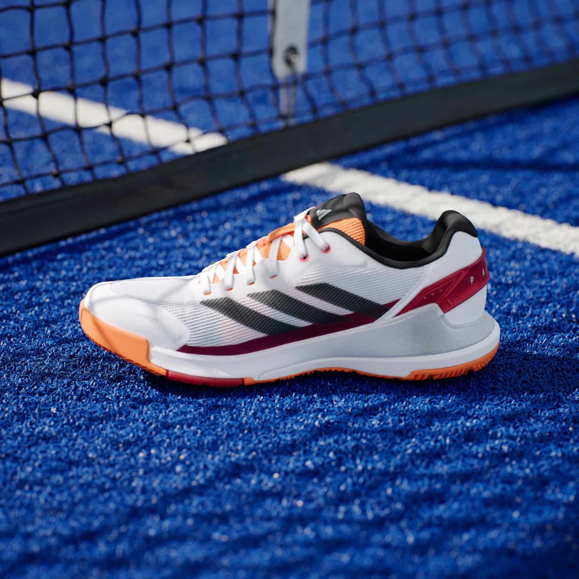 An adidas Crazyquick LS Mens Padel Shoe in white and orange with black stripes rests on blue turf beside a tennis net, offering superior multi-directional grip.