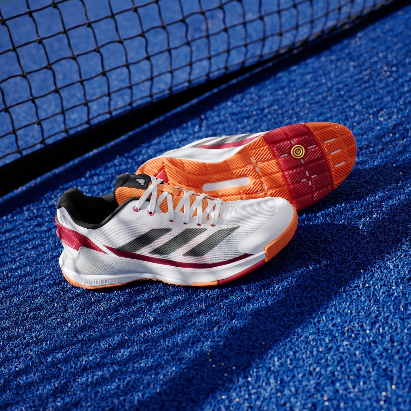 A pair of adidas Crazyquick LS Mens Padel Shoes in white with orange accents are set on a blue tennis court near the net.