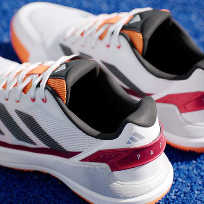 A close-up of adidas Crazyquick LS Mens Padel Shoes in White/Orange, featuring brand logos, laces, and a multi-directional grip sole, resting on a textured blue surface with red, gray, and orange accents.