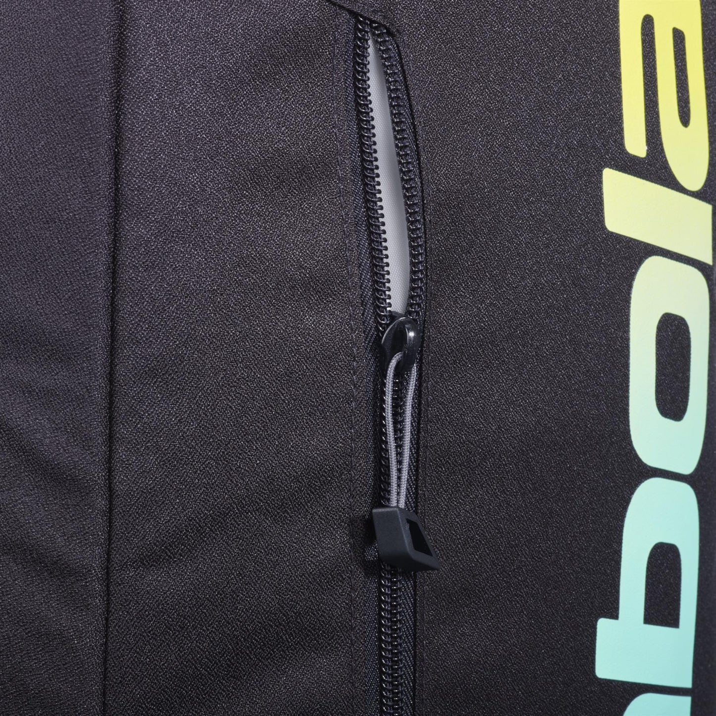 Close-up of the black zippered pocket on the Babolat Court Lite Padel Backpack - Black / Multi, showing part of the "Babolat" logo in a yellow-green gradient and a peek at the dedicated racket compartment on the right.