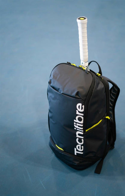 The Tecnifibre Reform Padel Backpack - Black by Tecnifibre, shown with yellow accents and a racket handle visible, stands ready on a blue court—a stylish, durable choice for carrying your padel gear.
