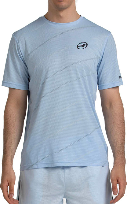 A person wears the Bullpadel Casinas Men's Padel T-Shirt in Glacier Blue, featuring a black circular logo, subtle diagonal lines, and Quickerdry technology for comfort.