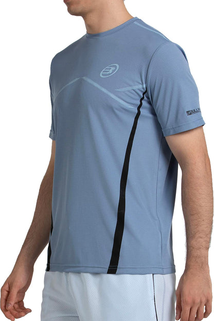 A man wearing the Bullpadel Cabore Men's Padel T-Shirt in shadow blue, featuring black vertical stripes, small logos on chest and sleeve, and Quickerdry technology. Shown from shoulders to hips. Brand: Bullpadel.