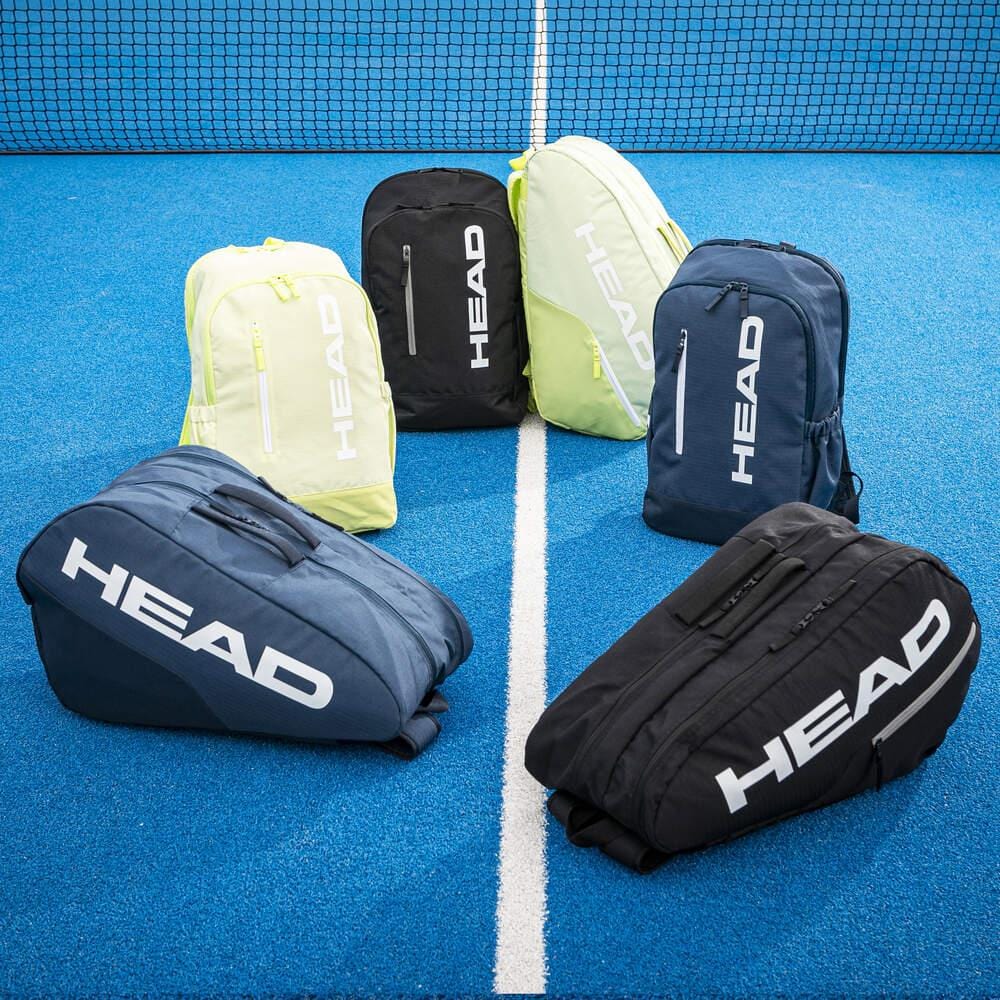 Six HEAD tennis bags, including the new HEAD BASE Padel Backpack 17L - Navy with a spacious racket compartment, are arranged in a semicircle on a bright blue tennis court with a white center line and net in the background.