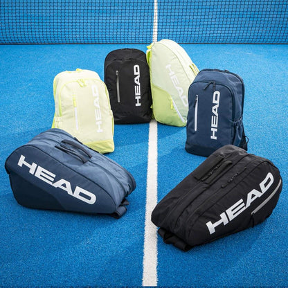 Six HEAD tennis bags, including the new HEAD BASE Padel Backpack 17L - Navy with a spacious racket compartment, are arranged in a semicircle on a bright blue tennis court with a white center line and net in the background.