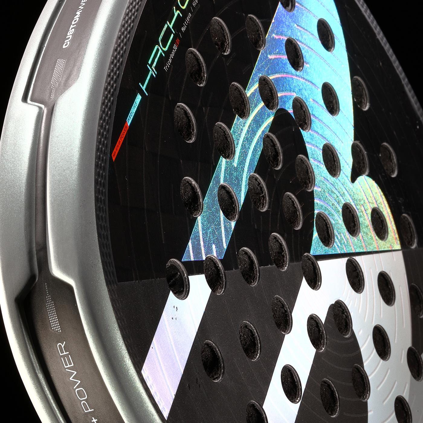 Close-up of the Bullpadel HACK 04 2026 Padel Racket featuring a metallic edge, circular perforations, and a colorful holographic pattern on its black surface. "HACK" and "POWER" are partially visible on the Bullpadel frame.