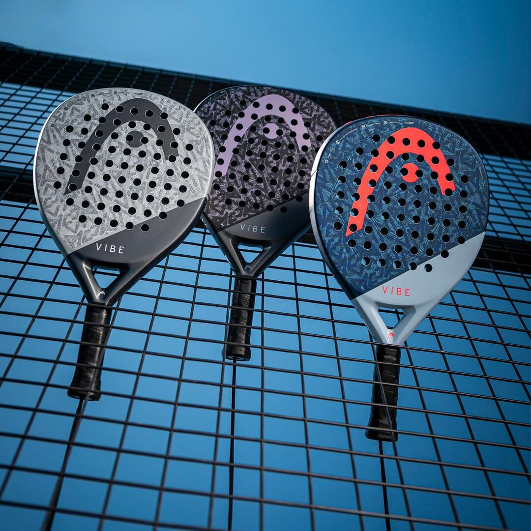 Three teardrop-shaped HEAD Vibe 2025 Grey / Black padel rackets with bold perforations hang on a wire fence against a blue sky, showcasing the dynamic design of the HEAD Vibe series.