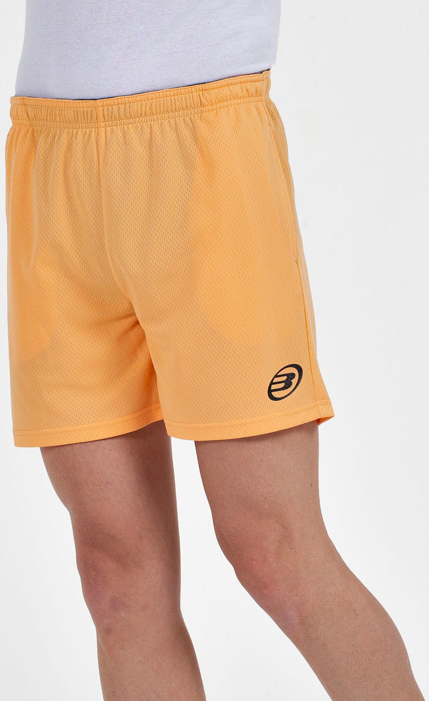 A man in Bullpadel Campaza Mens Padel Shorts - Orange, showcasing an elastic waistband and a small black Bullpadel logo on the left leg, is paired with a white shirt against a plain white background.