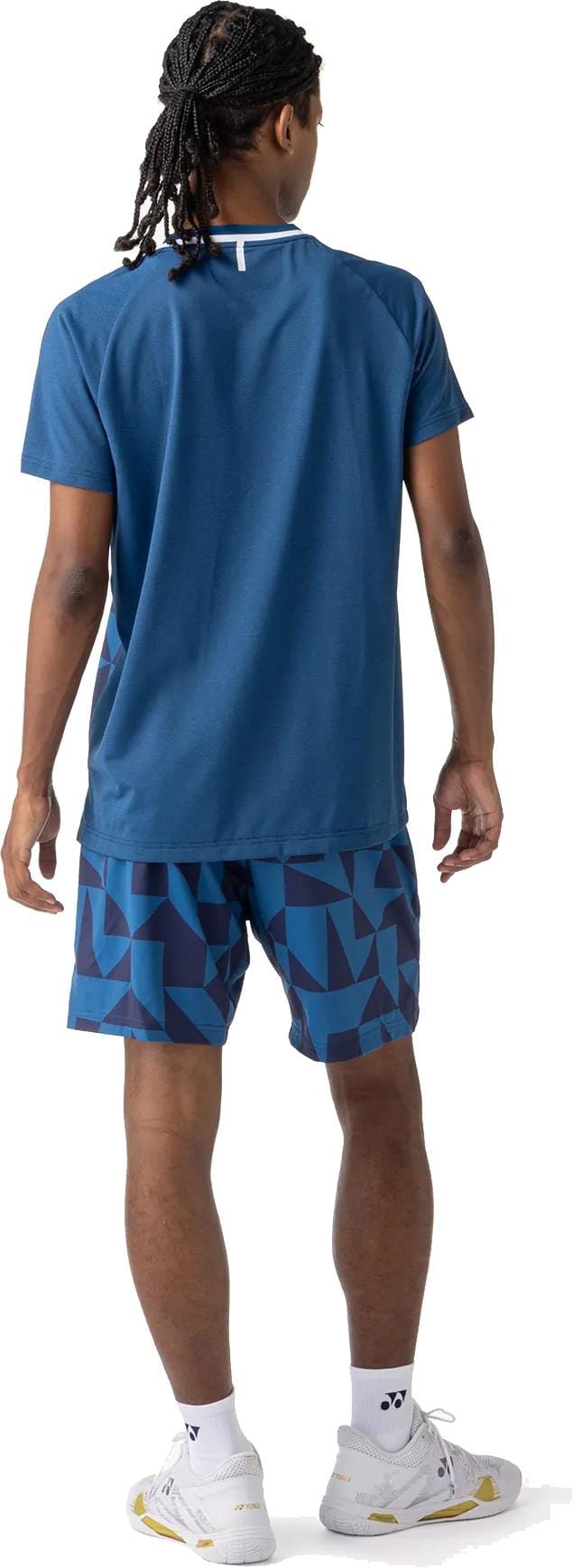 A person with braided hair, facing away, wears a blue t-shirt, Yonex 15240YX Knit Mens Padel Shorts in Indigo Blue by Yonex, white socks, and white sneakers.