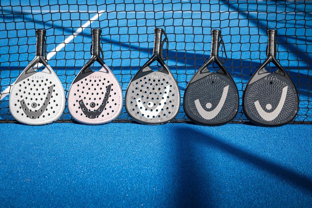 Five HEAD Speed One X 2025 Padel Rackets by HEAD in white, pink, light gray, dark gray, and black are arranged against a black net on a bright blue court. Sunlight highlights each racket featuring Auxetic 2.0 technology.