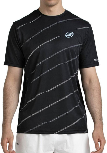 A man wears the Bullpadel Casinas Men's Padel T-Shirt in black, featuring diagonal white stripes and a small circular logo, made from lightweight polyester with Quickerdry technology, paired with white shorts against a plain background.